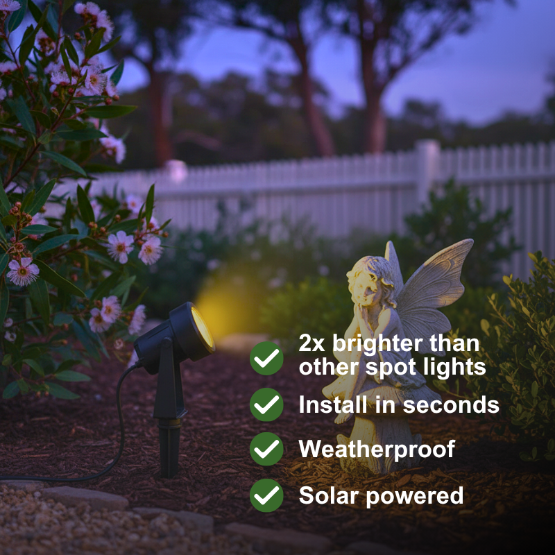 A black solar-powered garden spotlight shown in a backyard setting at dusk, casting a warm yellow light onto the mulch and plants. A small angel garden statue stands nearby, with promotional text listing benefits: 2x brighter than other spot lights, install in seconds, weatherproof, and solar powered.