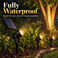 Three black solar-powered landscape spotlights installed in a garden at night during rain, casting warm yellow light onto plants and a tree trunk. Large text at the top reads 'Fully Waterproof' and 'Built for rain, dew & Aussie weather'.